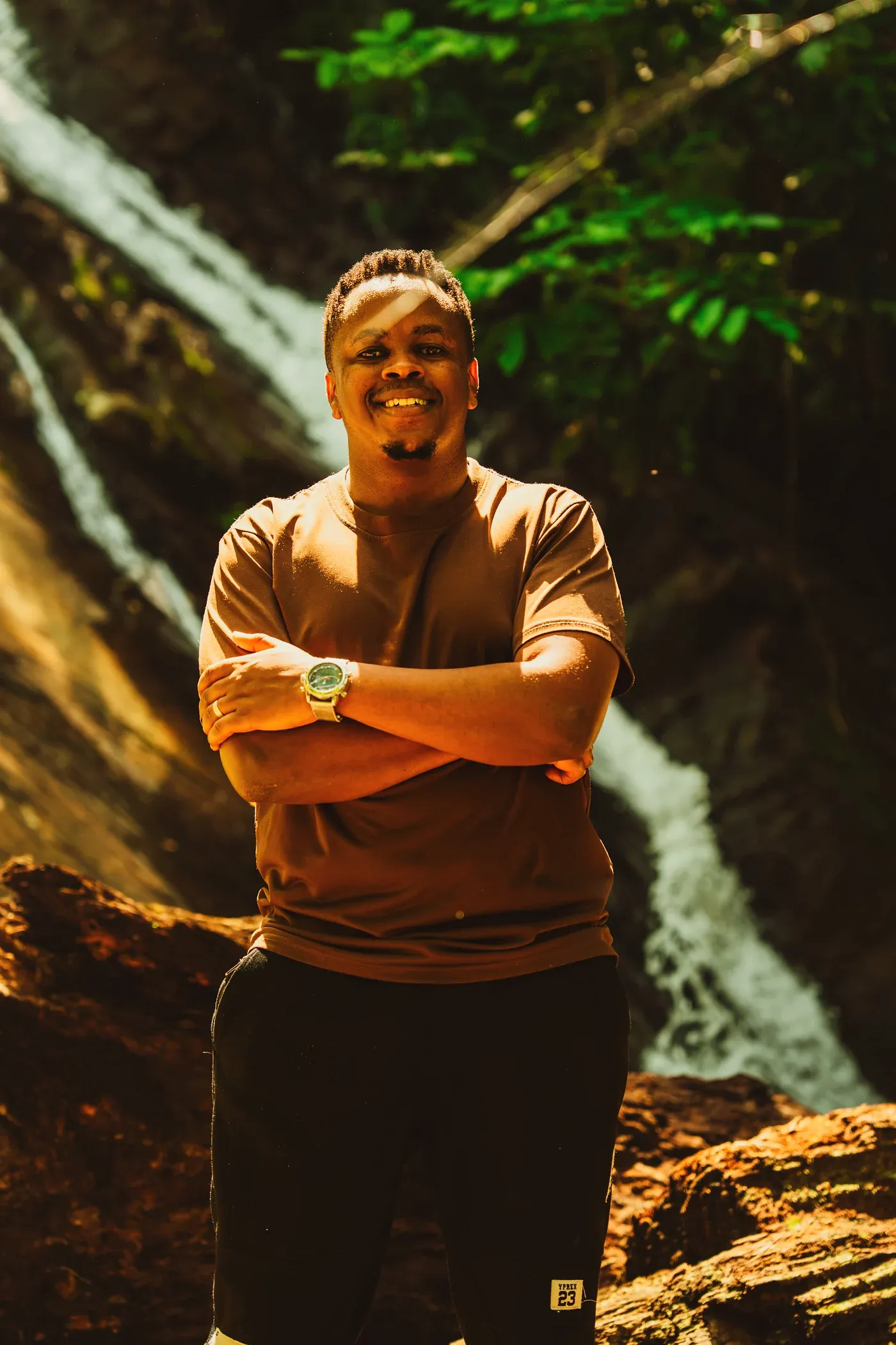 Jordan Mtui smiling with arms crossed in front of a forest waterfall