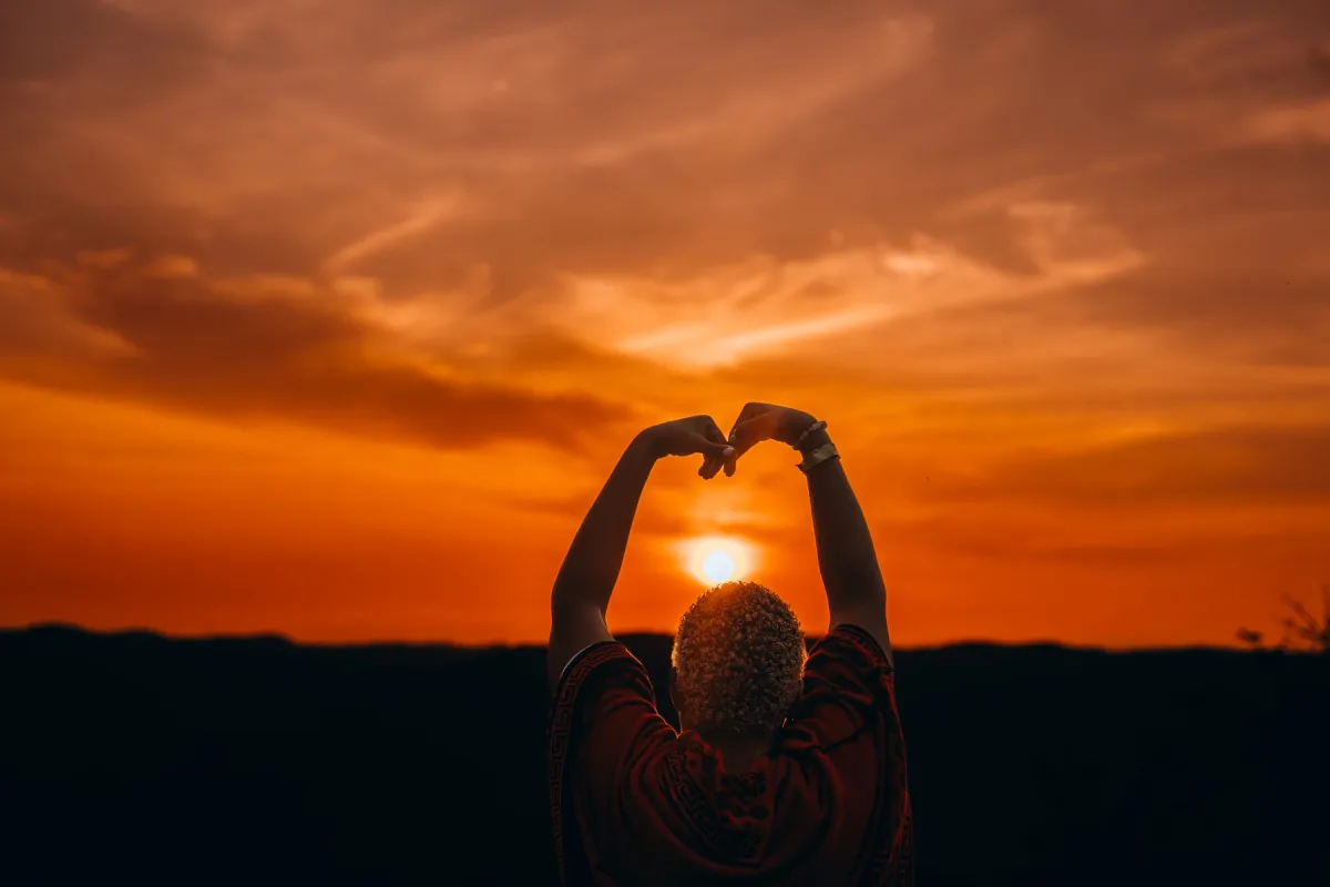 A person making a heart shape with their hands against an orange Magoroto sunset